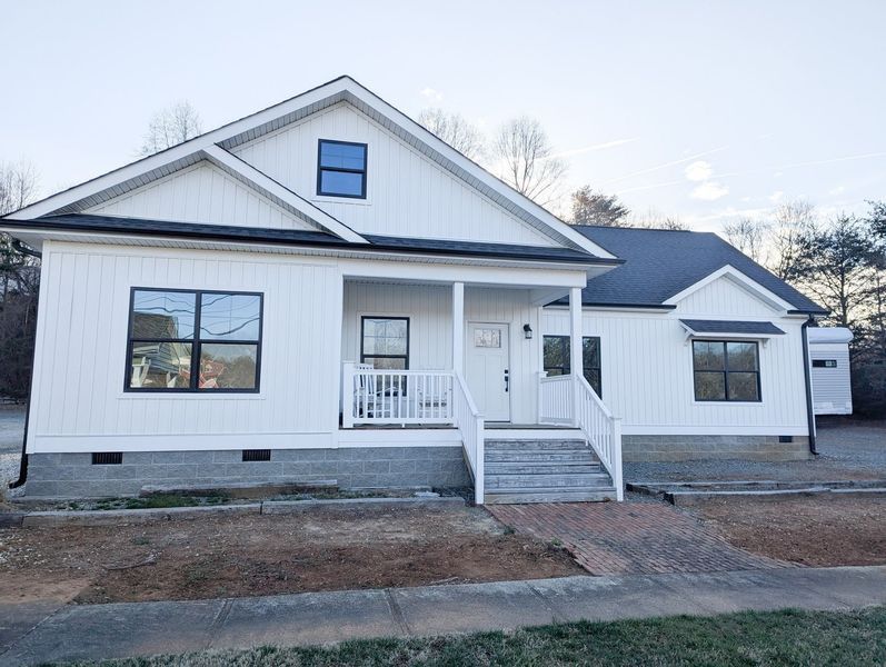 A newly constructed, white farmhouse-style home with vertical siding, a front porch, and a dark shingled roof.