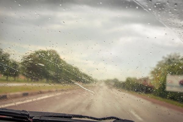 Rainy road view through a car windshield with wipers and wet pavement under a cloudy sky