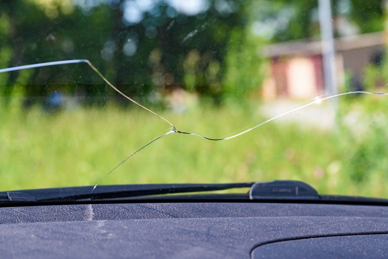 Cracked windshield seen from inside a car, with blurred greenery and a building outside.