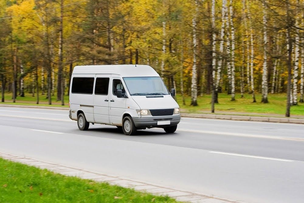 White van driving on a road beside a park with yellow autumn trees