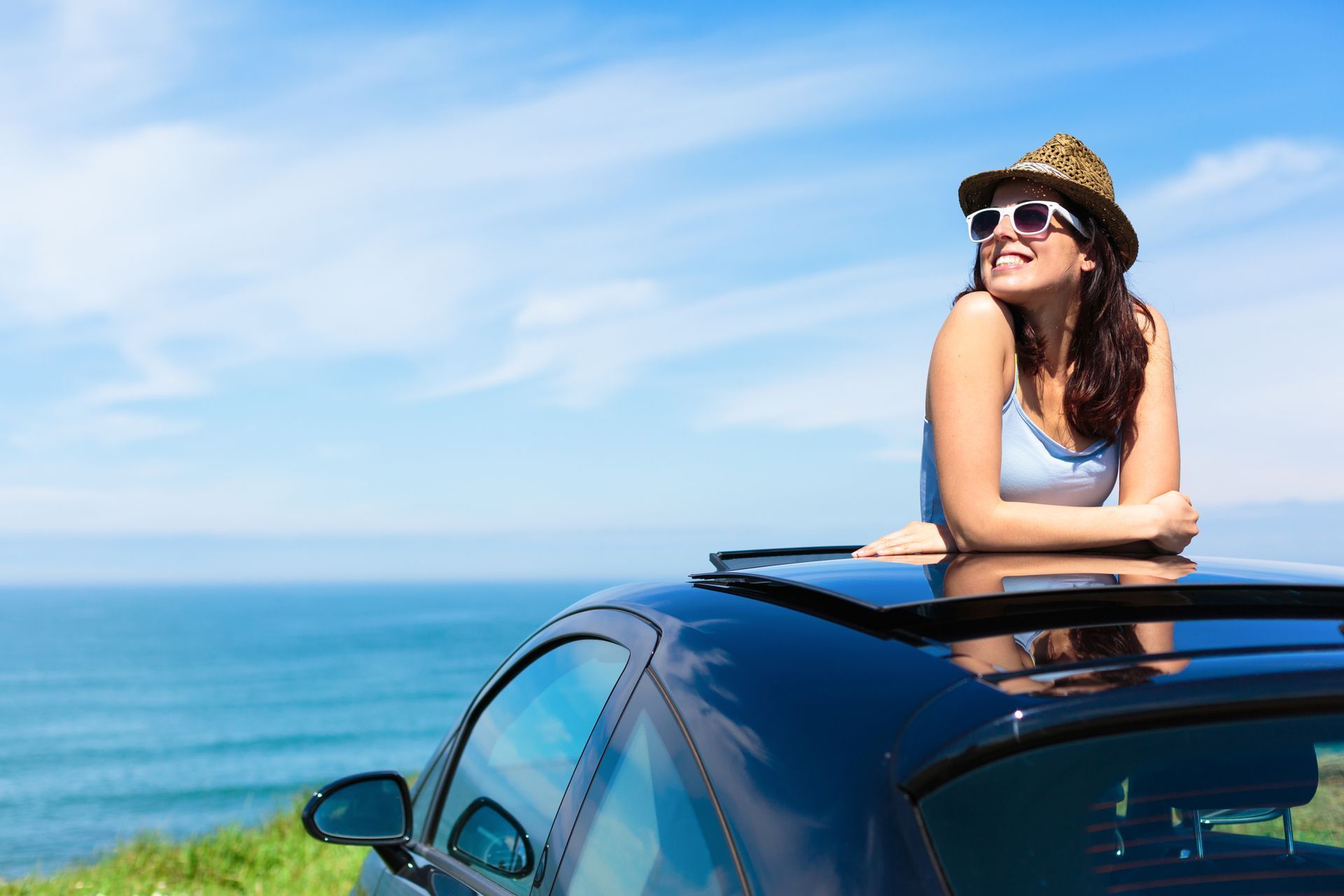 Woman leaning on a car roof by the ocean, wearing sunglasses and a hat under a blue sky.