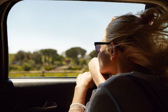 Person leaning out a car window, resting on their arm, with a sunny landscape outside