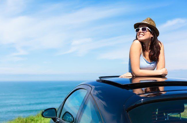Woman leaning on a car roof by the ocean, wearing a hat and sunglasses, under a blue sky