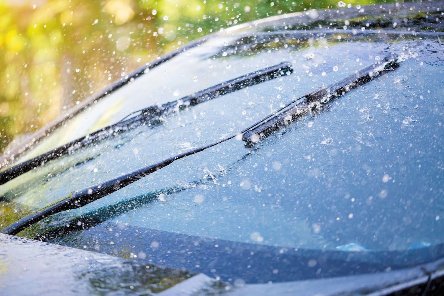 Windshield wipers clearing rain from a car windshield with droplets and blue sky reflections