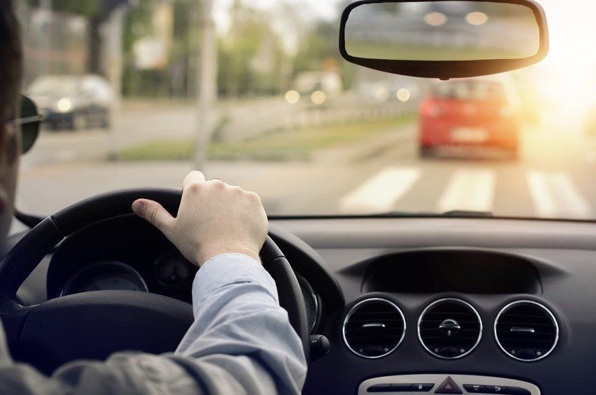 Driver holding steering wheel inside car, driving on a sunlit road with another car ahead.