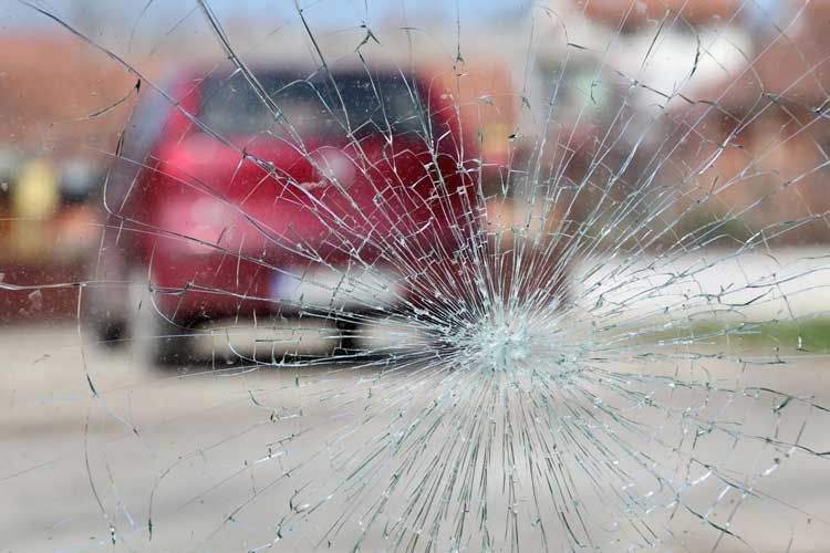 Cracked windshield with shattered glass, blurred red car in the background.