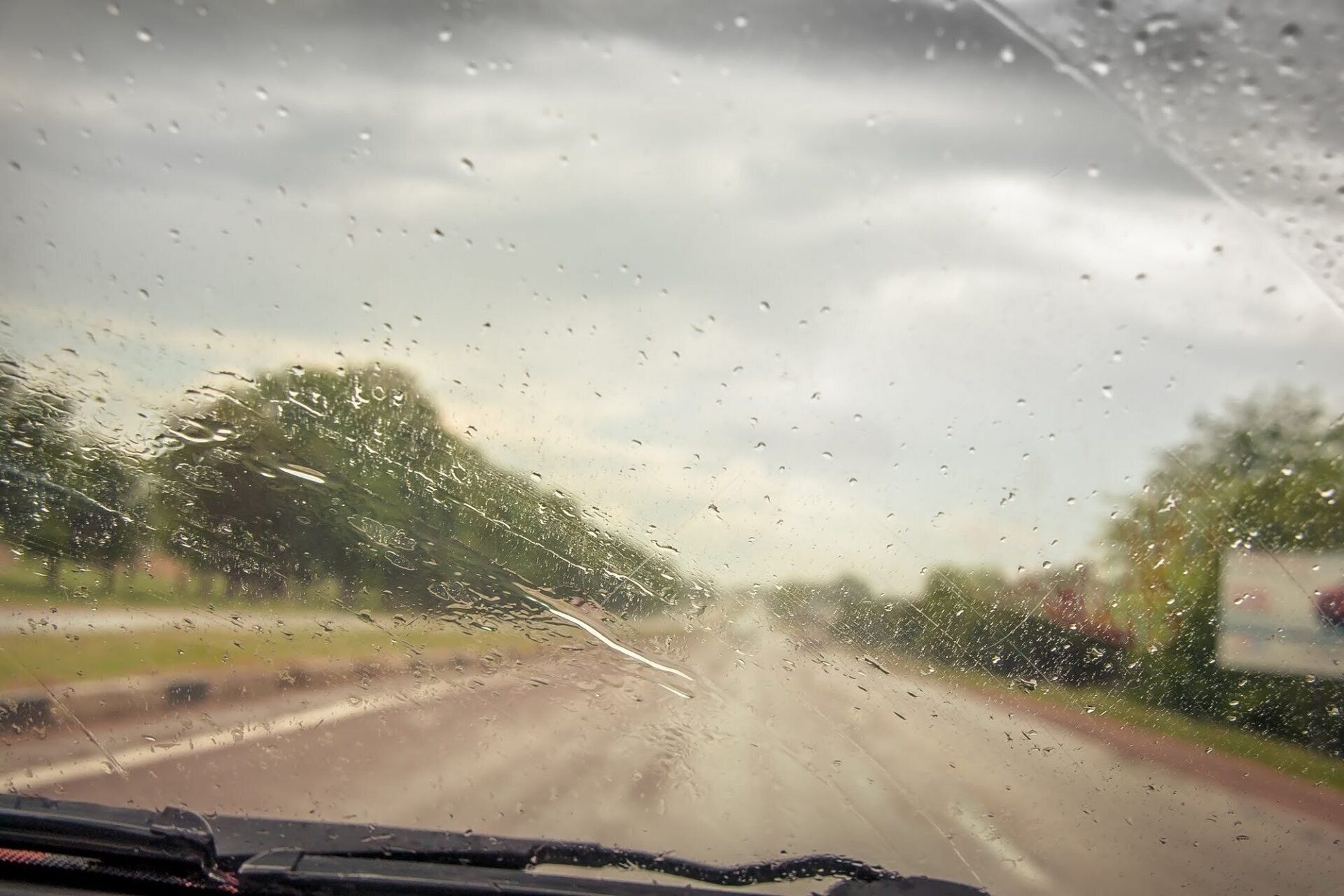 Rainy road seen through a car windshield with raindrops and blurred trees under a cloudy sky