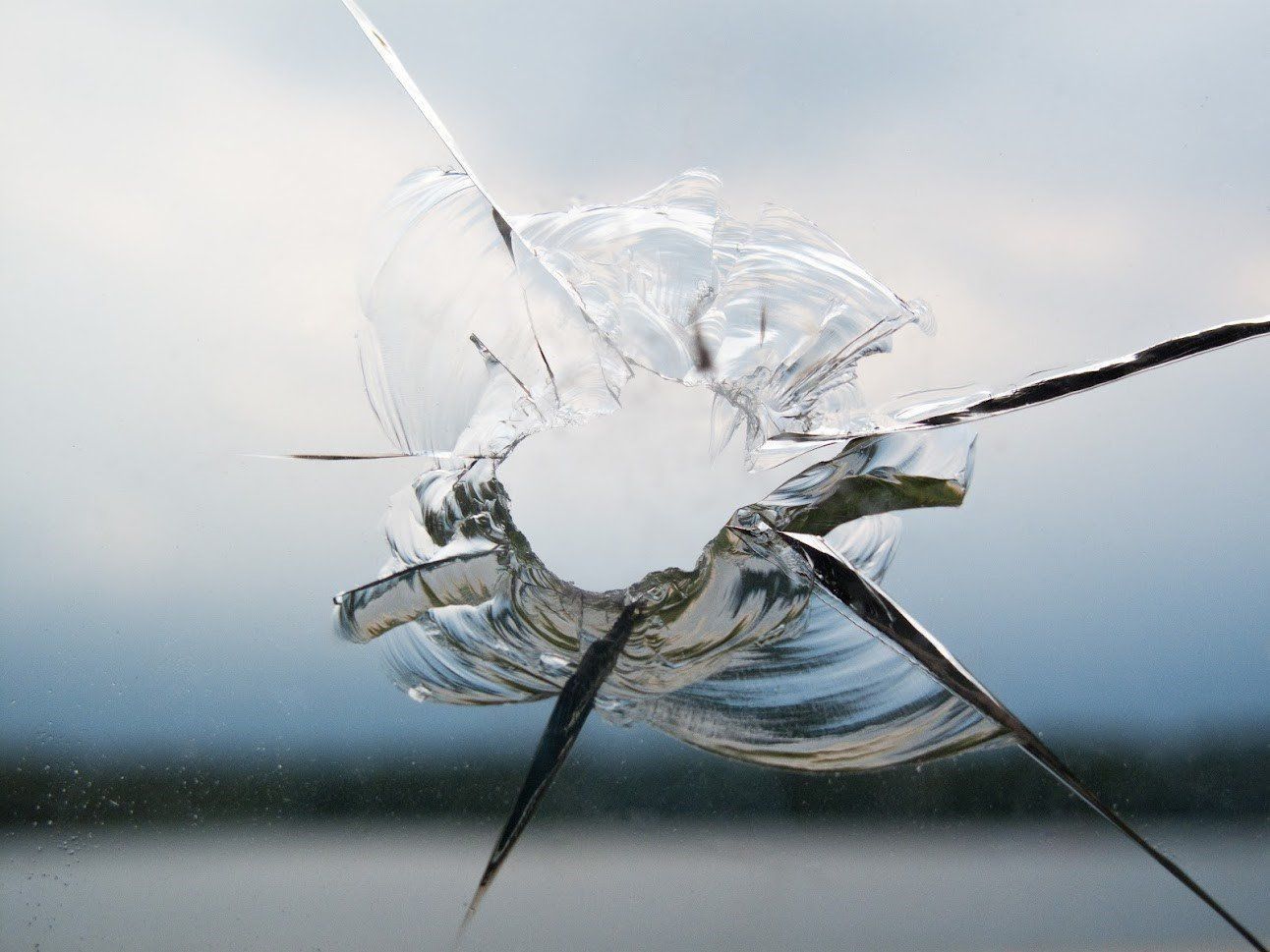 Glass bullet hole in a windshield with cracked shards against a blurred sky and horizon.