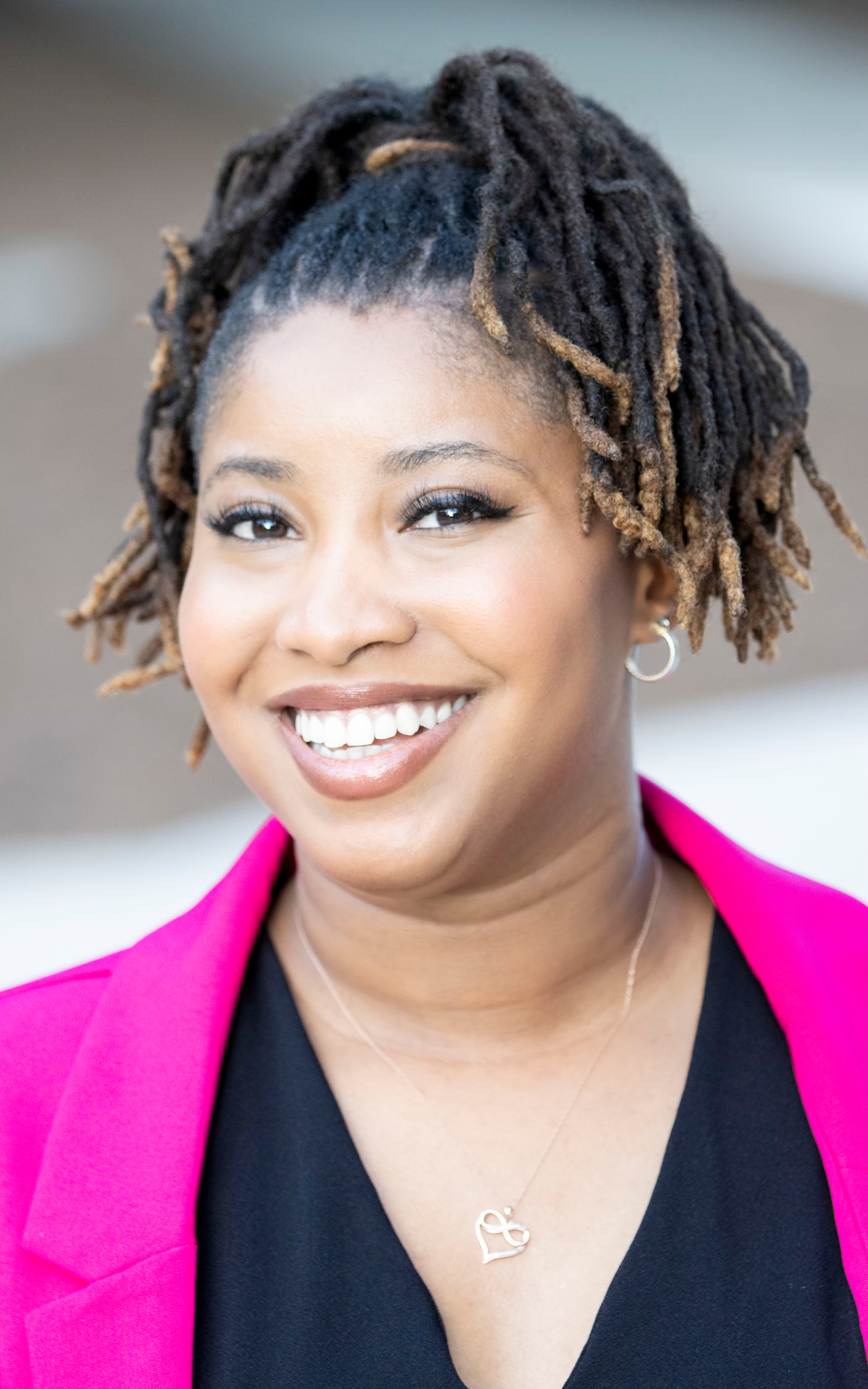 Attorney Alycia Vail Taylor smiling, wearing a black top and bright pink blazer outdoors.