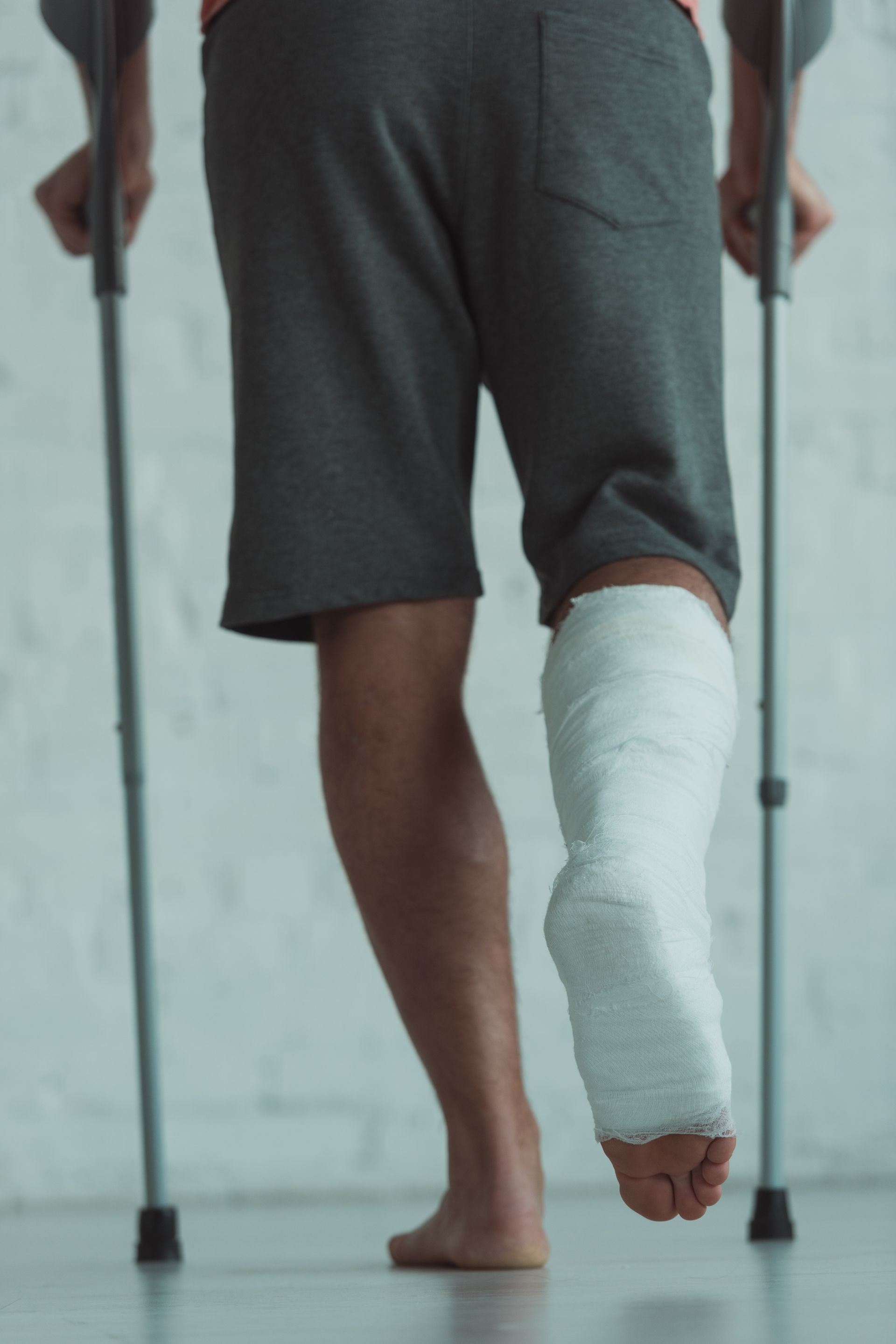 Person walking on crutches with a leg in a cast; gray shorts, indoors.