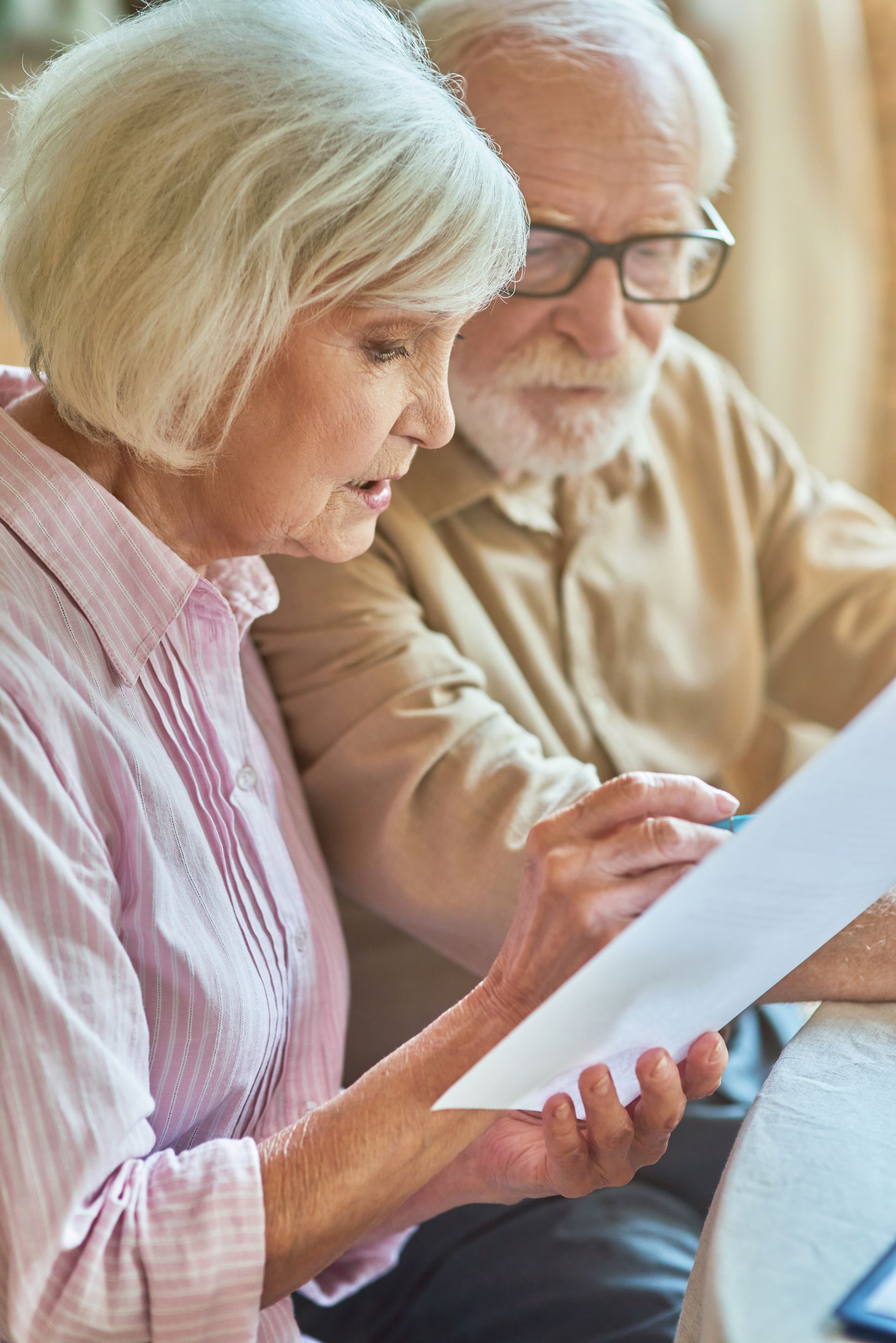 Elderly couple reviewing documents, looking concerned; indoors. Woman in pink shirt, man with glasses.