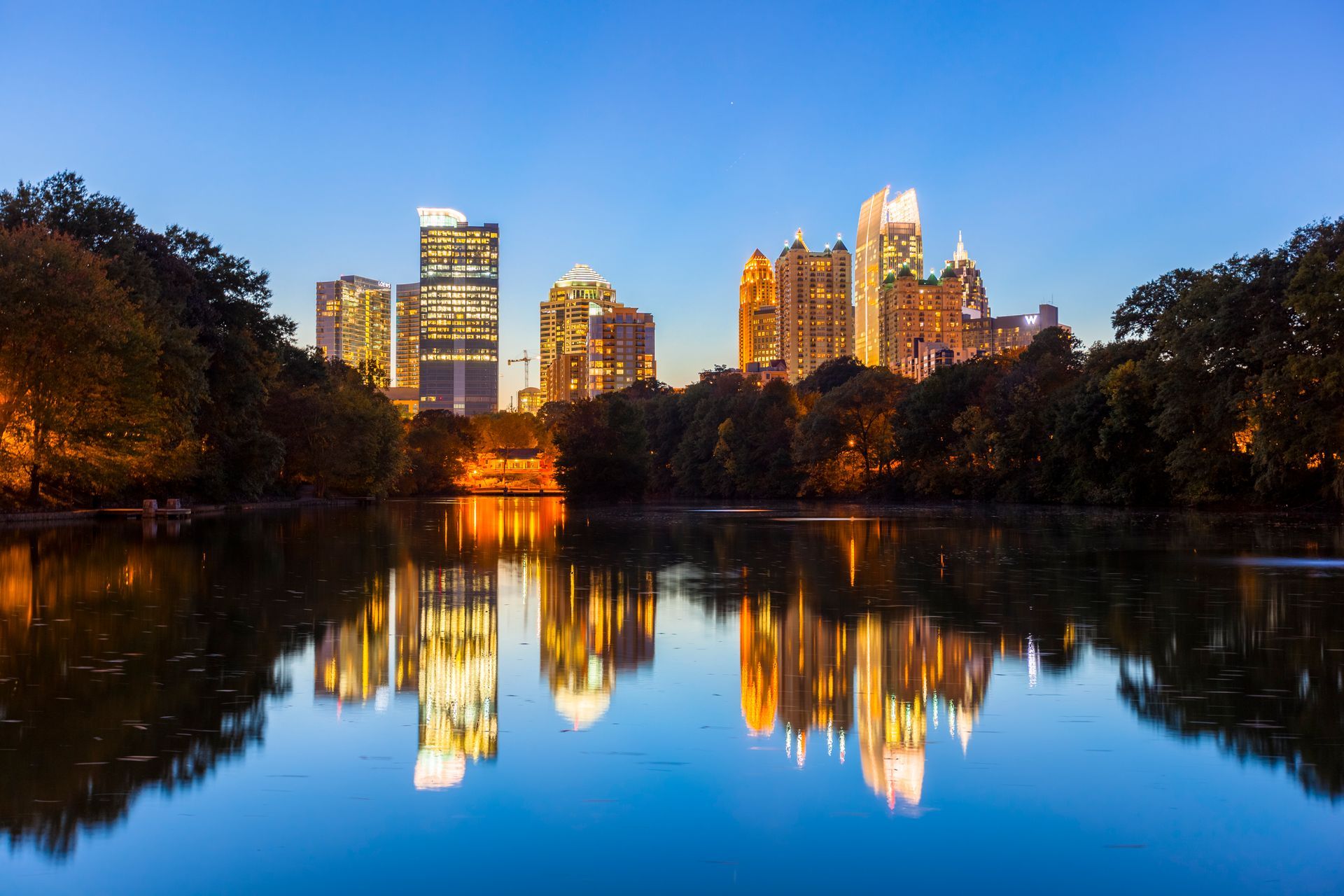 City skyline reflecting in a calm lake at twilight; illuminated buildings, trees along shore, blue sky.