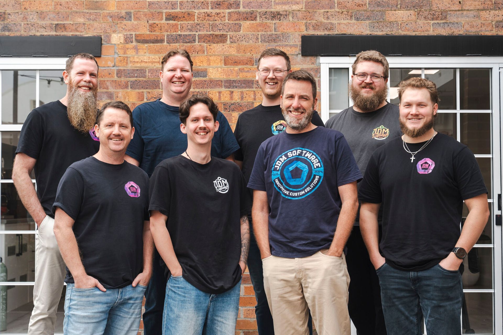Seven people posing together outdoors in front of a brick wall, wearing black T-shirts and smiling.