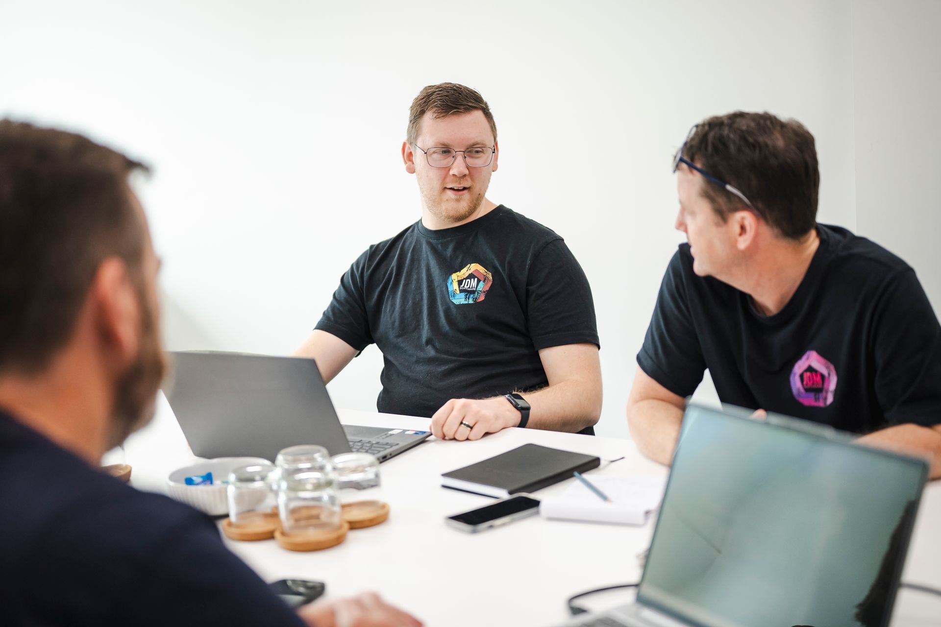 Three people in a meeting around laptops, discussing documents at a white table.