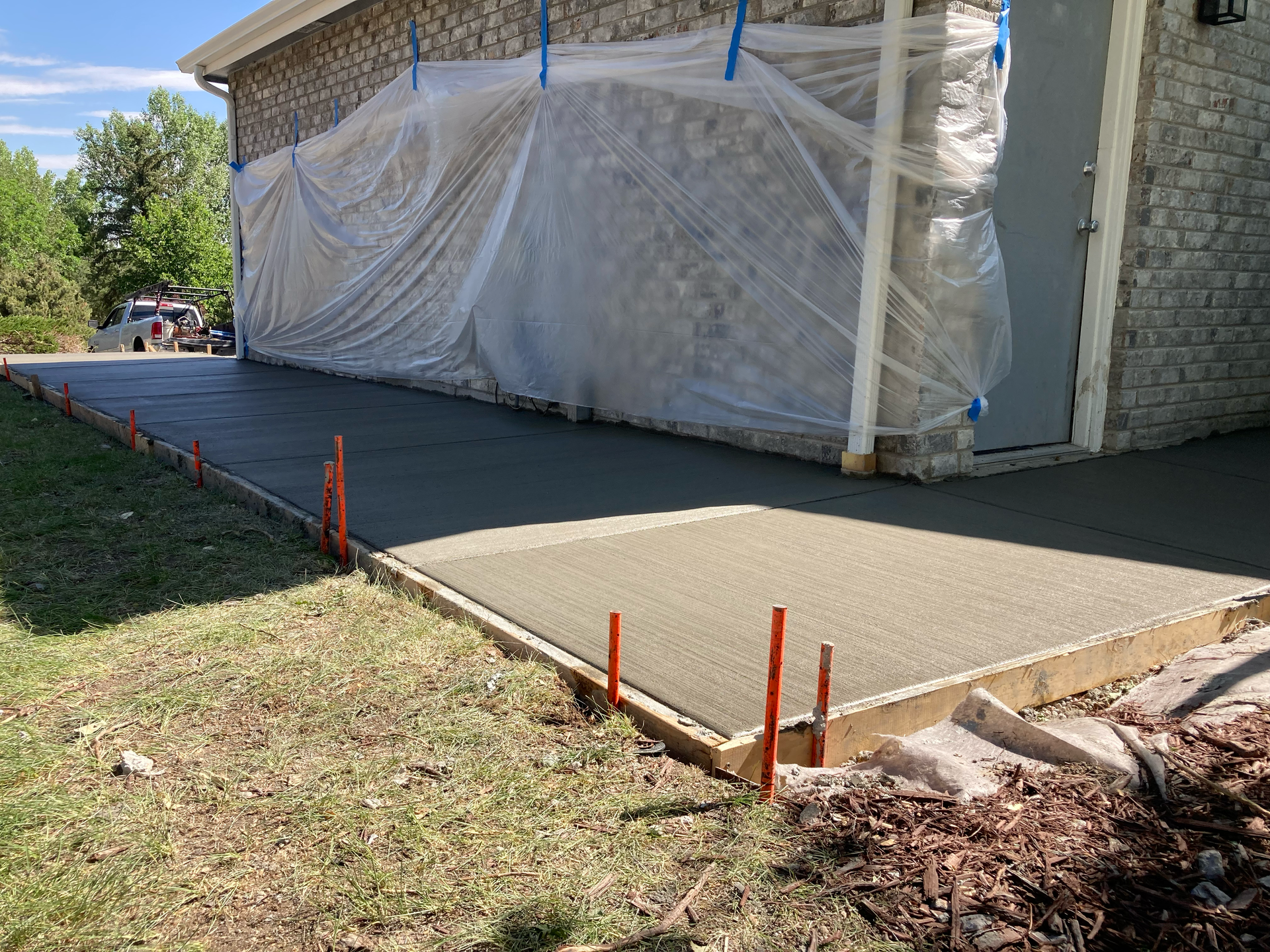 Newly poured side patio slab with orange stakes marking the forms, house draped in plastic