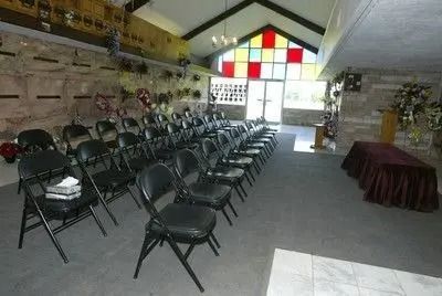 Indoor event hall with rows of folding chairs facing a bright stained-glass window and a covered table on the right