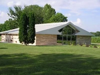 Low stone building with a peaked roof and large windows, surrounded by green lawn and trees