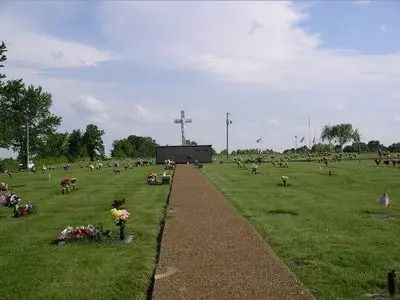 Cemetery with a central path leading to a large cross, surrounded by graves and flowers under a cloudy sky