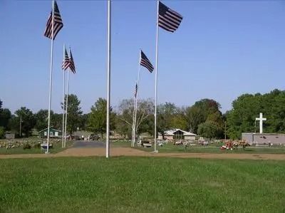 Four American flags flying above a small memorial with a white cross and green lawn.