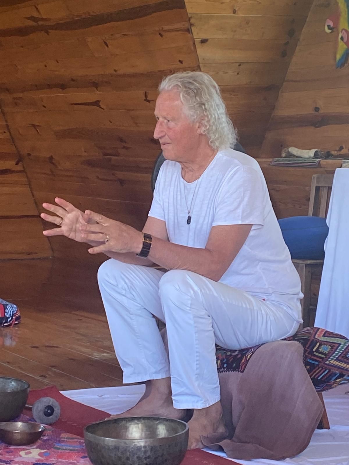 Mann mit weißen Haaren in weißer Kleidung, gestikuliert im Sitzen, Innenraum.
