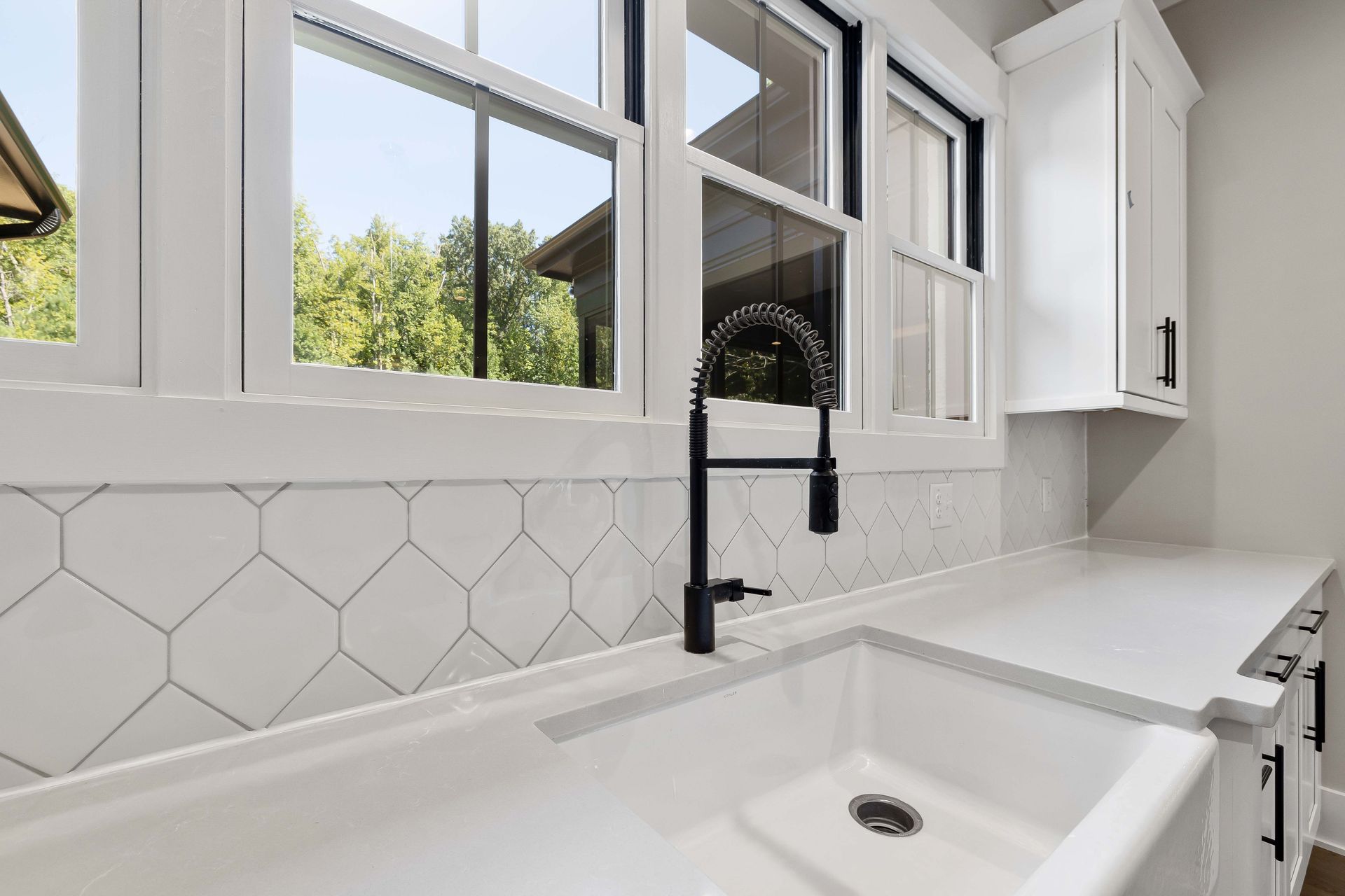 Residential kitchen sink in front of gray backsplash and three windows with a view of a tree line.  