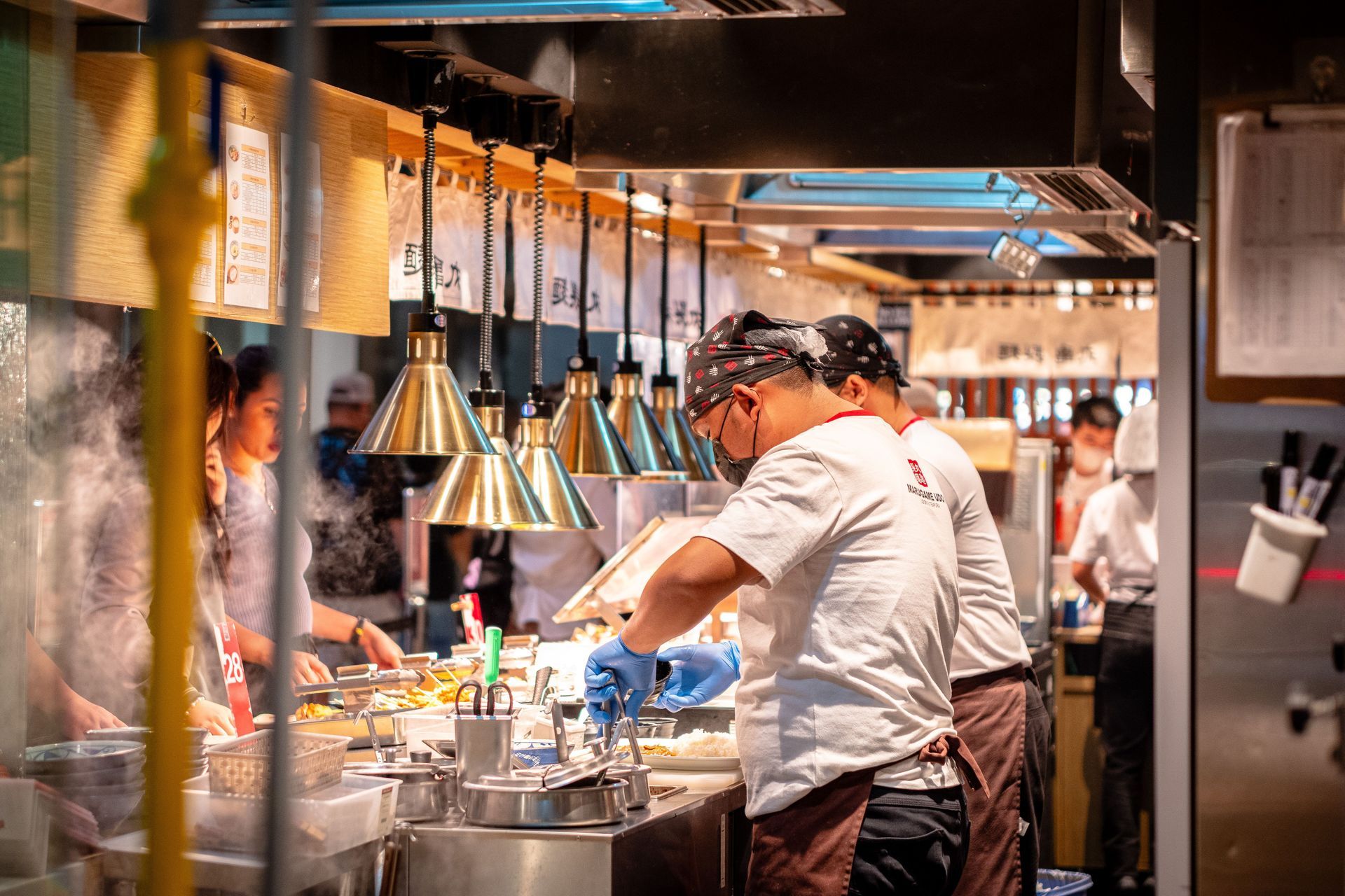Workers wearing masks at an Asian restaurant preparing food for the customers forming a line in front of the kitchen counter.