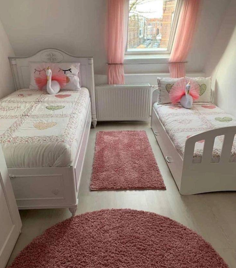 Two white beds with pink bedding, a rug, and pink curtains in a light-filled room.