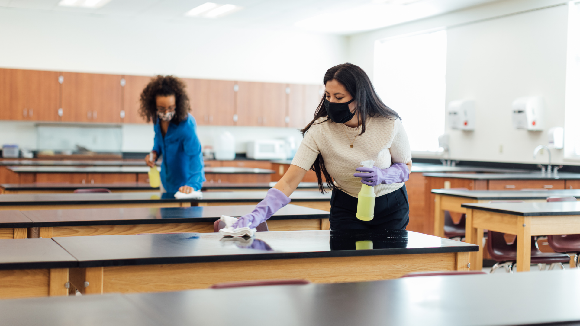Two people wearing masks and gloves sanitize classroom desks.
