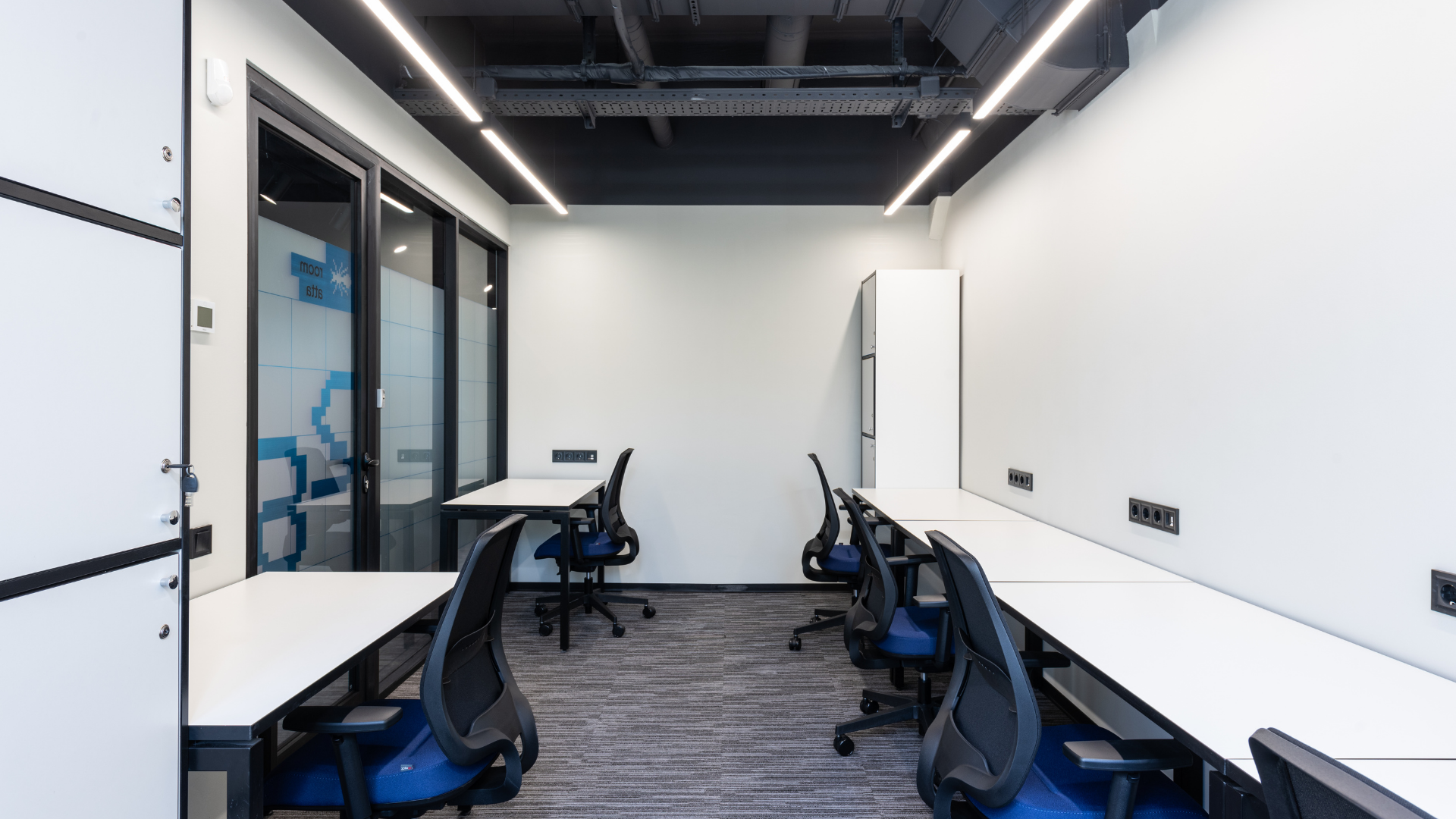 Office room with white desks, blue chairs, and glass wall, under bright lights.