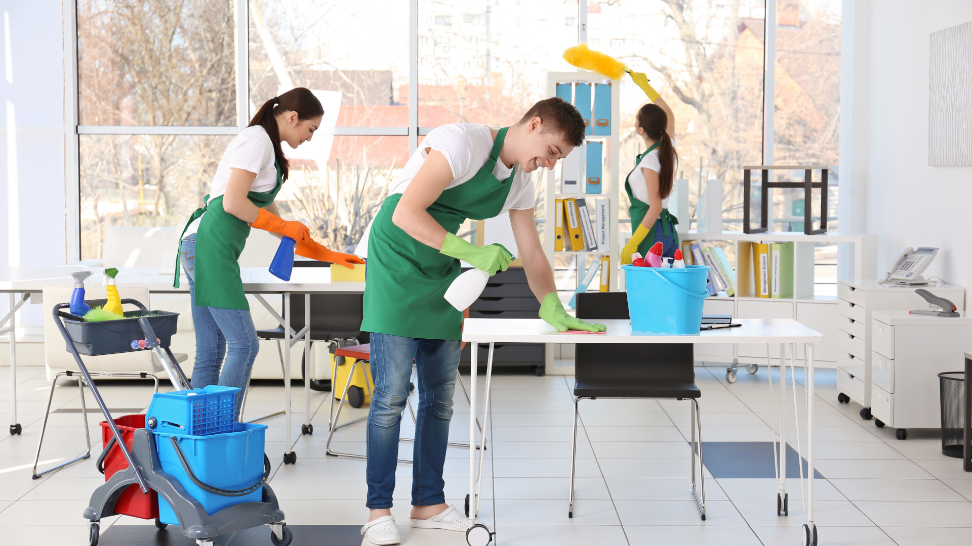 Three people cleaning an office. Two are wiping desks, one dusting. Bright, open room with windows.