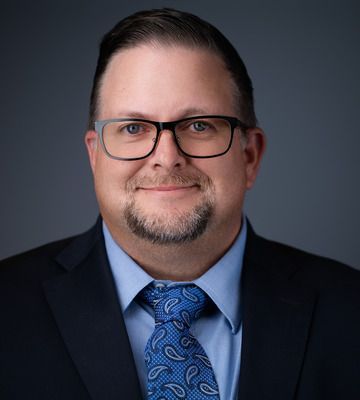 Studio headshot of a man in glasses wearing a navy suit, light blue shirt, and patterned blue tie.