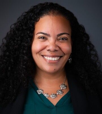 Smiling person with curly hair wearing a teal top and silver necklace against a dark background