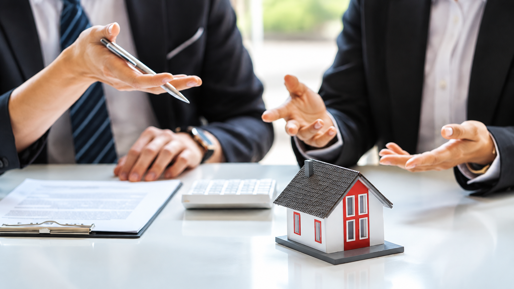 Hands discussing a house model on a desk with calculator and clipboard in an office setting