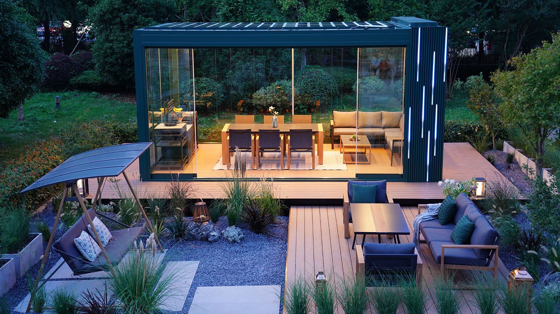 Modern backyard patio with a glass-walled lounge, wooden deck, and outdoor seating surrounded by greenery.