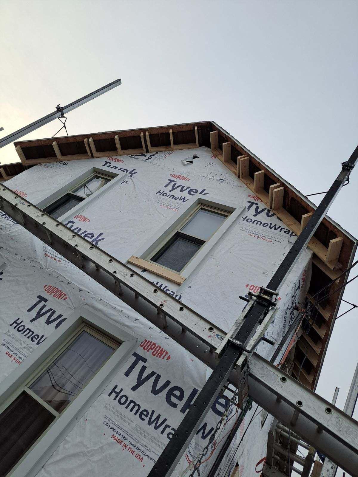 House under construction wrapped in Tyvek, viewed from below with scaffolding and roof framing visible