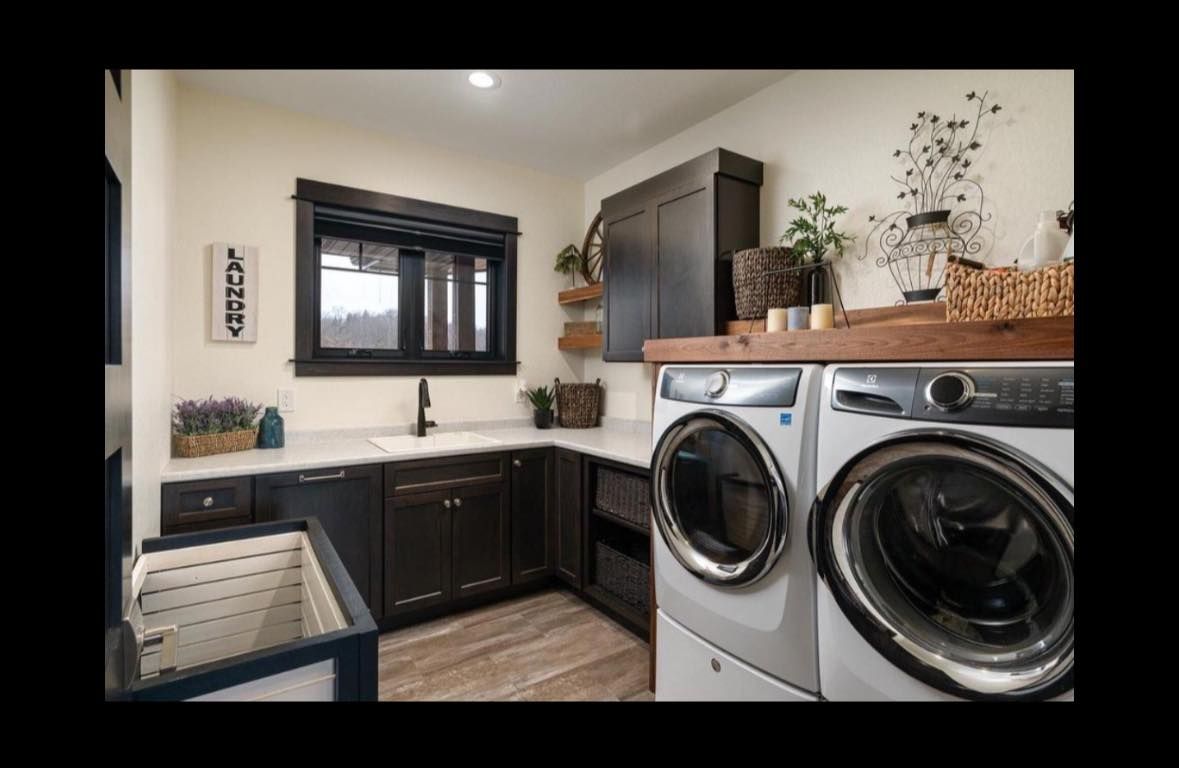 Modern laundry room with front-loading washer and dryer, dark cabinets, black window, and wood-look floor.