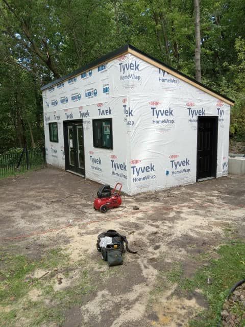Tyvek-wrapped small house under construction in a wooded lot, with a red mower in front.