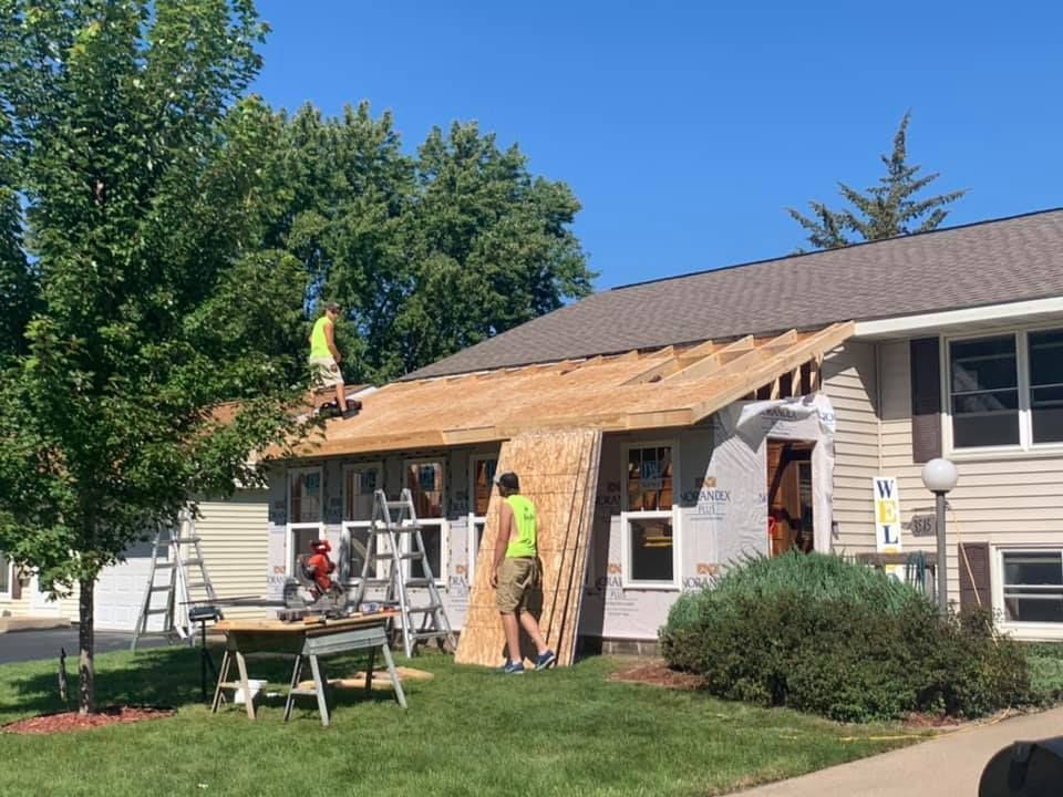 Workers installing shingles on a house roof with ladders and scaffolding in a sunny yard