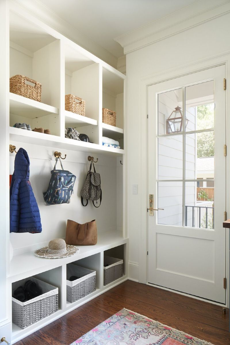 Bright entryway mudroom with white built-in cubbies, woven baskets, hanging coats, and a glass-paneled door.