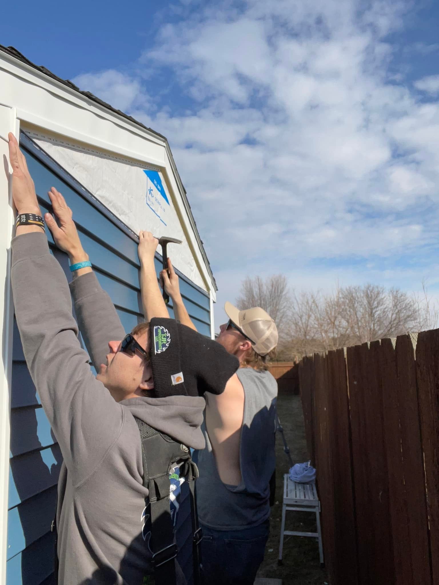 Two people on a ladder and one standing below reach up to install siding on a house exterior.