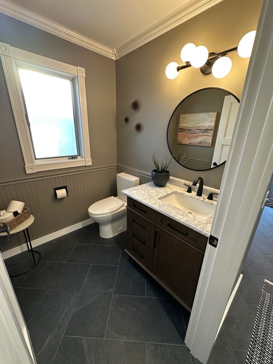 Small bathroom with dark tile floor, vanity sink, toilet, mirror, lights, and window.