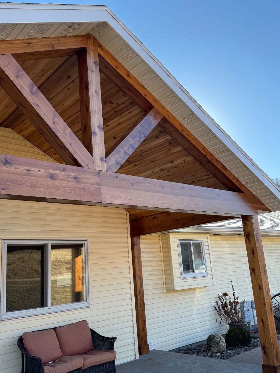 Covered porch with wooden timber trusses, beige siding, a window, and a bench under a sloped roof.