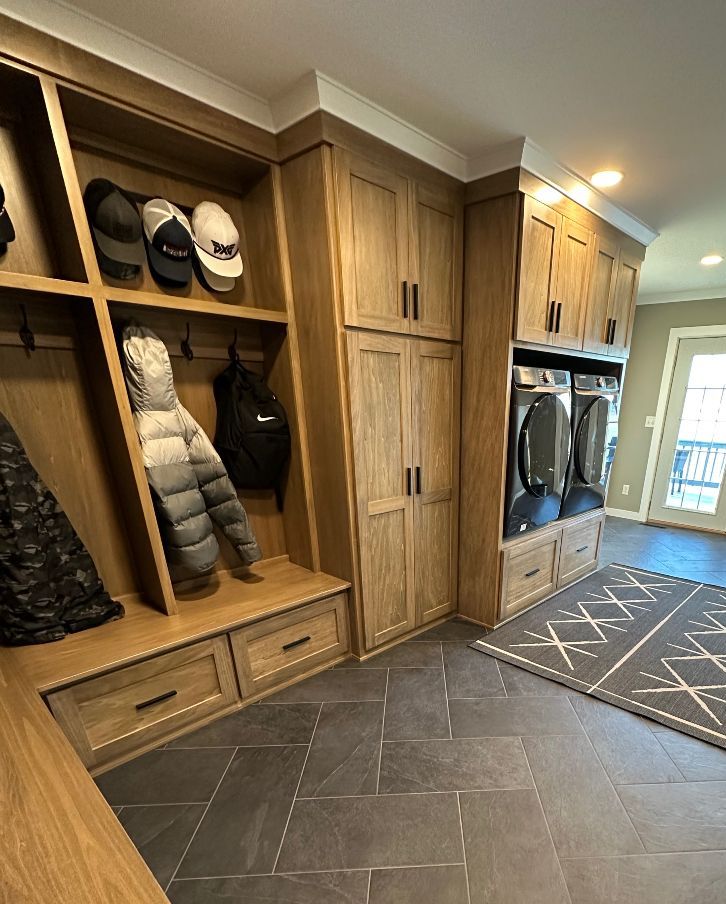 Built-in wood mudroom cabinets with coats, shoes, bags, and dark tile floor by a front door.