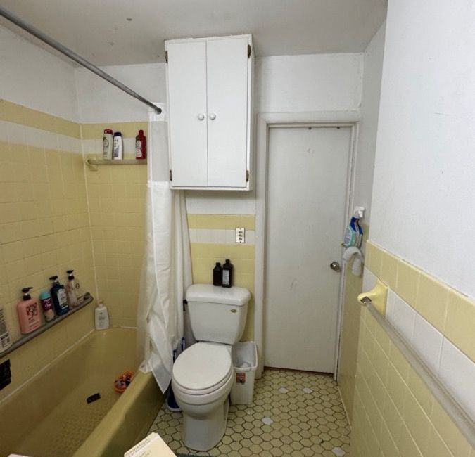 Small bathroom with toilet, yellow tile trim, wall cabinet, shower rod, and closed door.