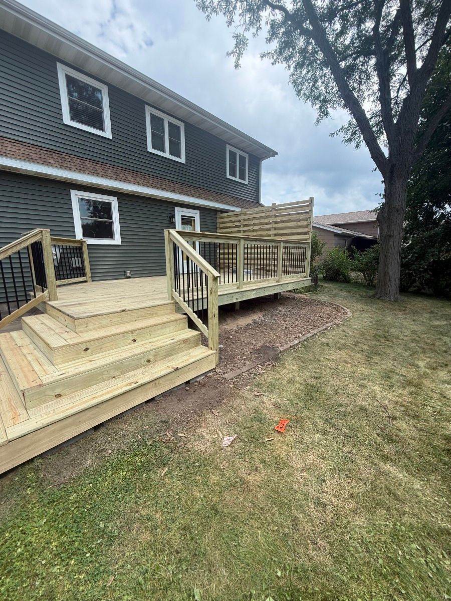 Wooden deck and stairs beside a dark gray house in a grassy yard with a large tree nearby