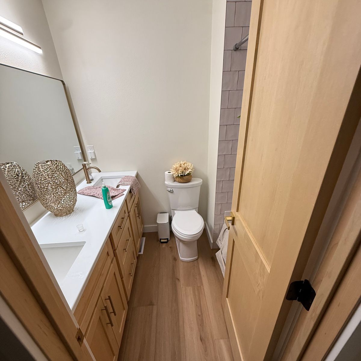 Narrow bathroom with white toilet, wood vanity, mirror, and light wood door partially open.
