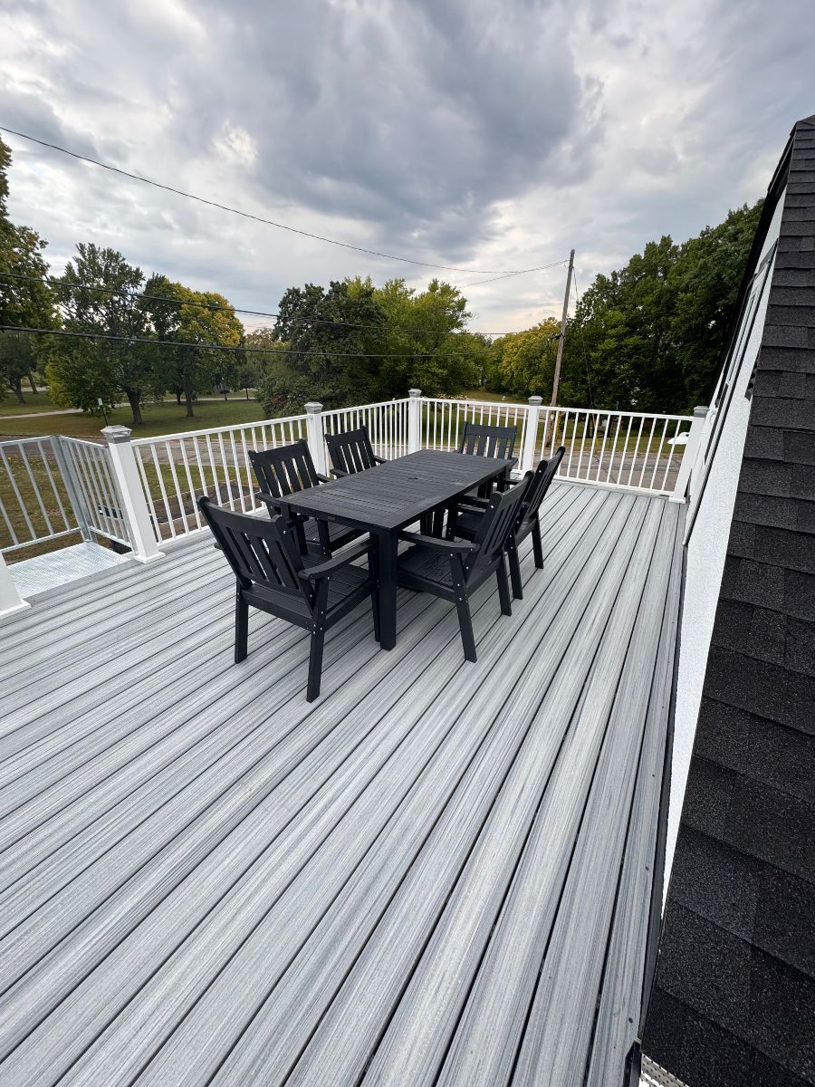 Outdoor deck with a black dining table and chairs, white railing, and cloudy sky beyond.