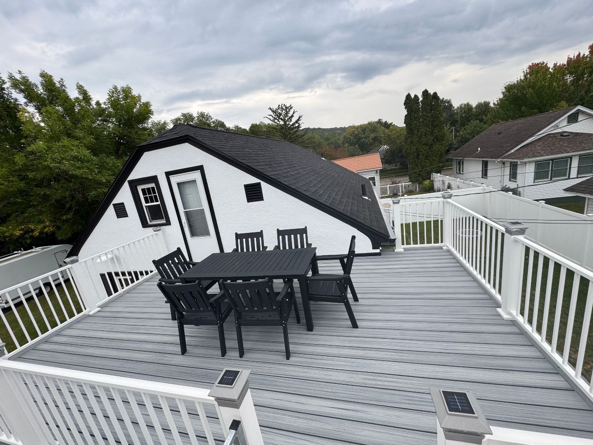 Rooftop deck with white railing, black patio table and chairs, and a small house under a cloudy sky