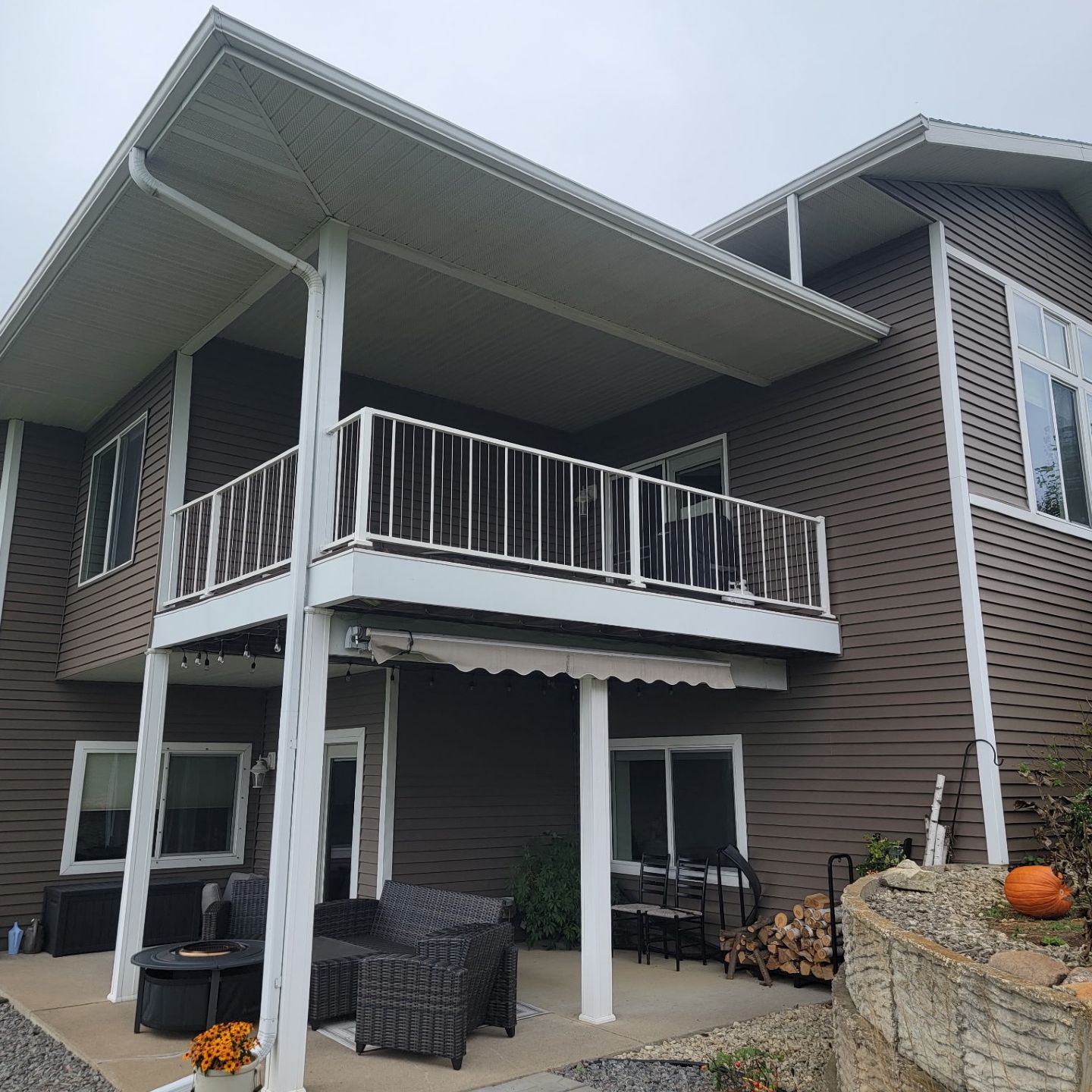 Two-story brick house with a covered balcony and patio furniture underneath, viewed from the yard.