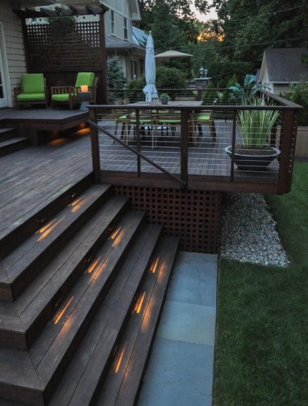 Backyard deck with lit steps, railing, patio seating, and a potted plant by a grassy yard